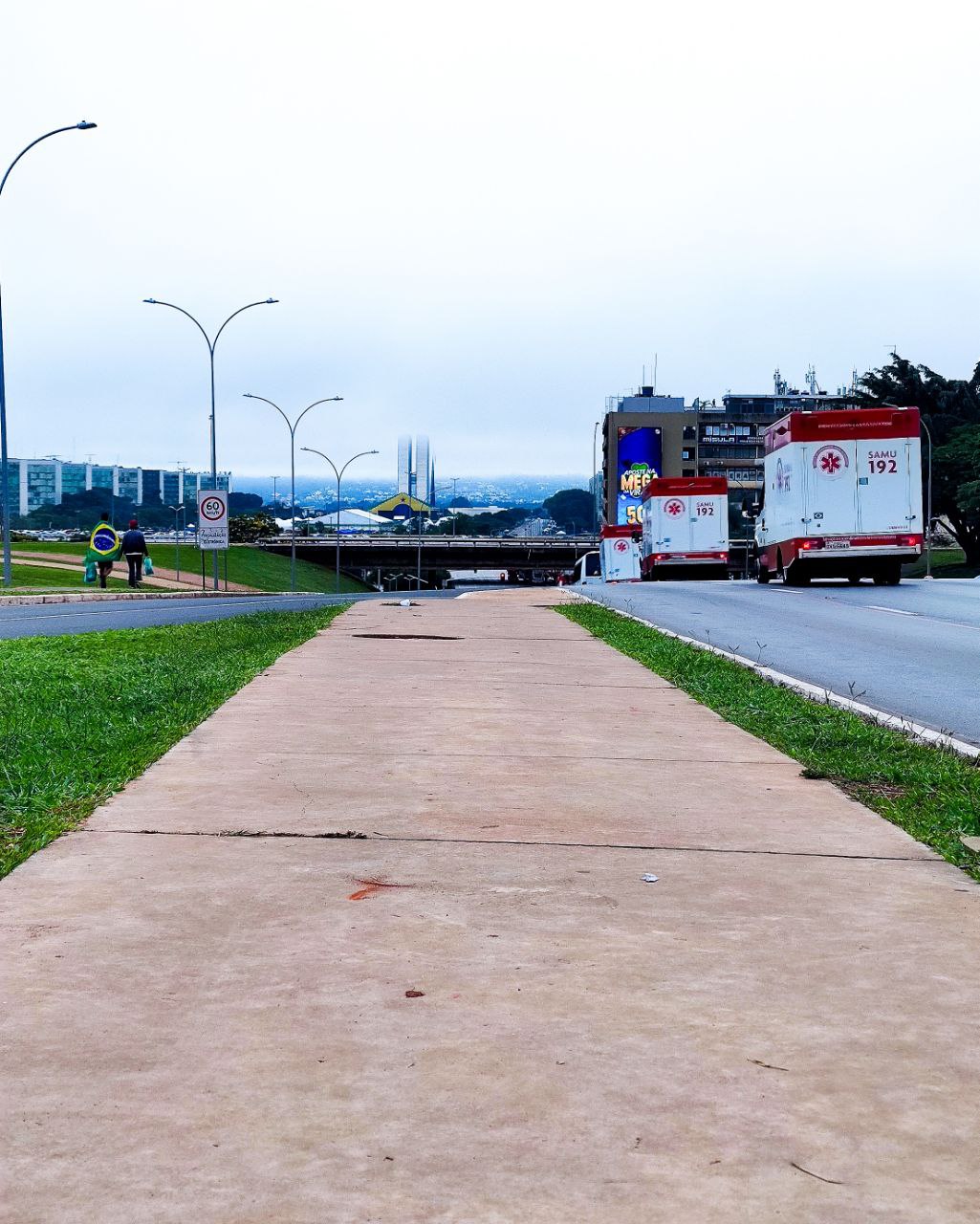 Brasília. Vista para o Eixo Monumental de Brasília. Pedestres caminham enrolados em bandeira nacional e duas ambulâncias transitam.