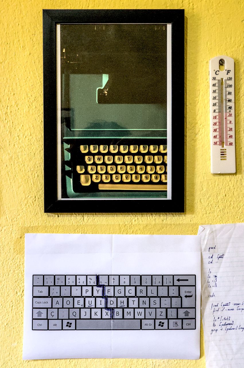 Convívio entre leiautes. Parede amarela com fotografia vertical emoldurada com máquina de escrever vista de cima, impressão de leiaute de teclado Dvorak e termômetro.