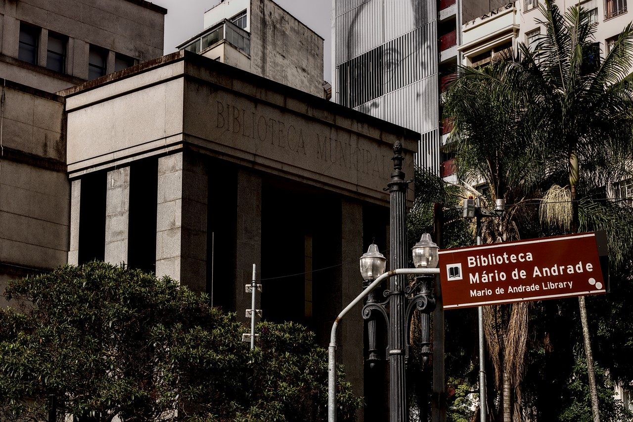 Fachada da Biblioteca Mário de Andrade, entrada da R. da Consolação. Pórtico cinzento de concreto. Ao fundo, retrato de Mário de Andrade em edifício. Num plano adiante, poste de luz da antiga Light Co.