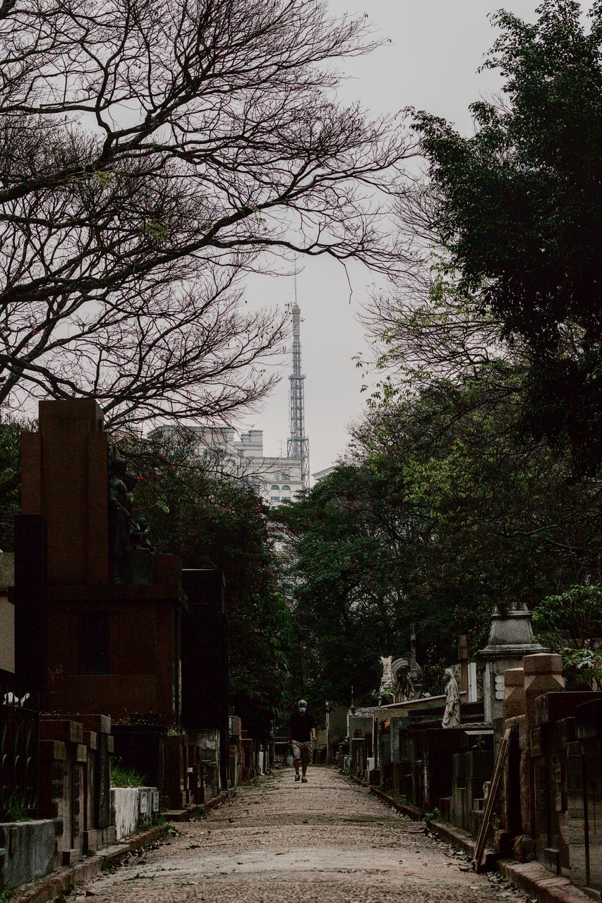 Homem portando máscara facial caminha entre túmulos; alta torre de rádio e TV da Band ao fundo