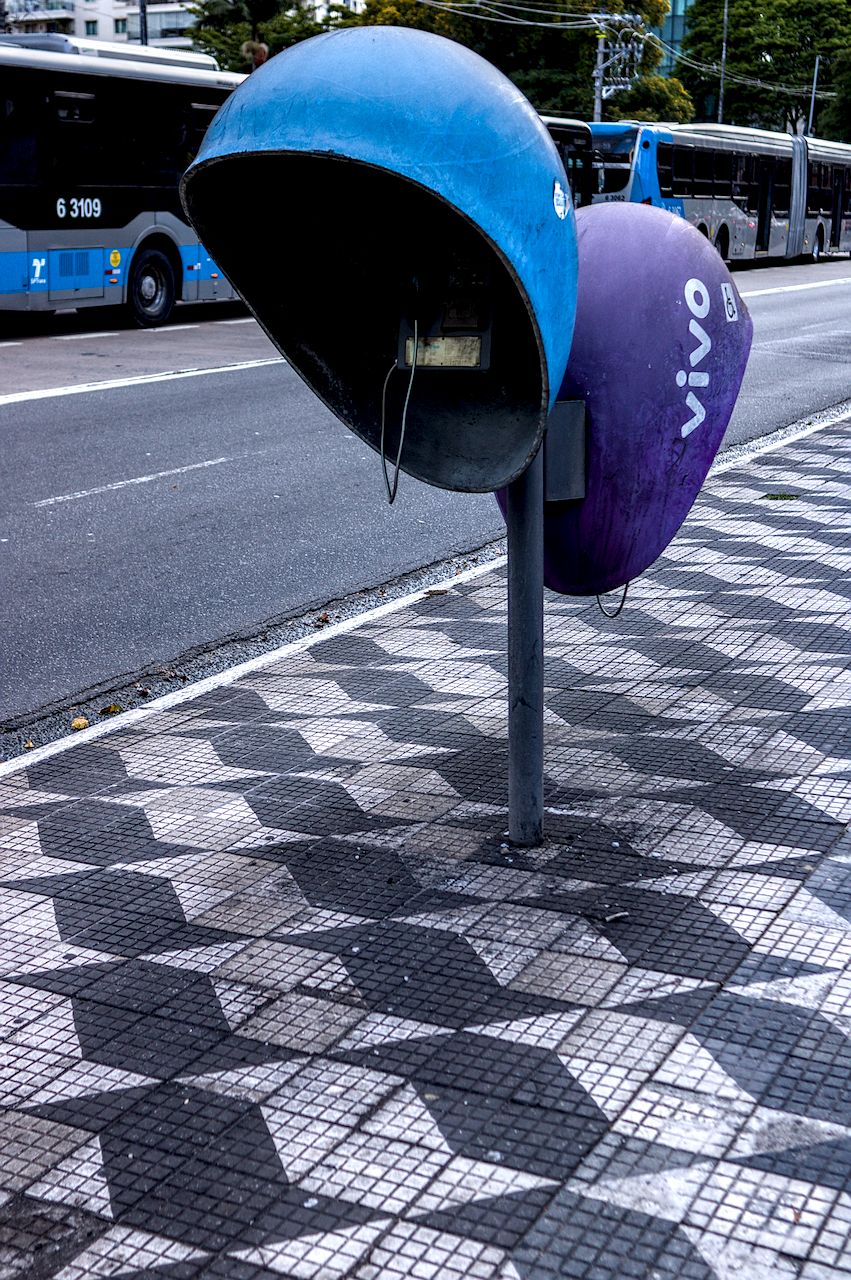 Orelhão e mosaico paulista. Dois orelhões azul e púrpura em calçada de mosaico paulista. Ao fundo e em desfoque, ônibus azul.