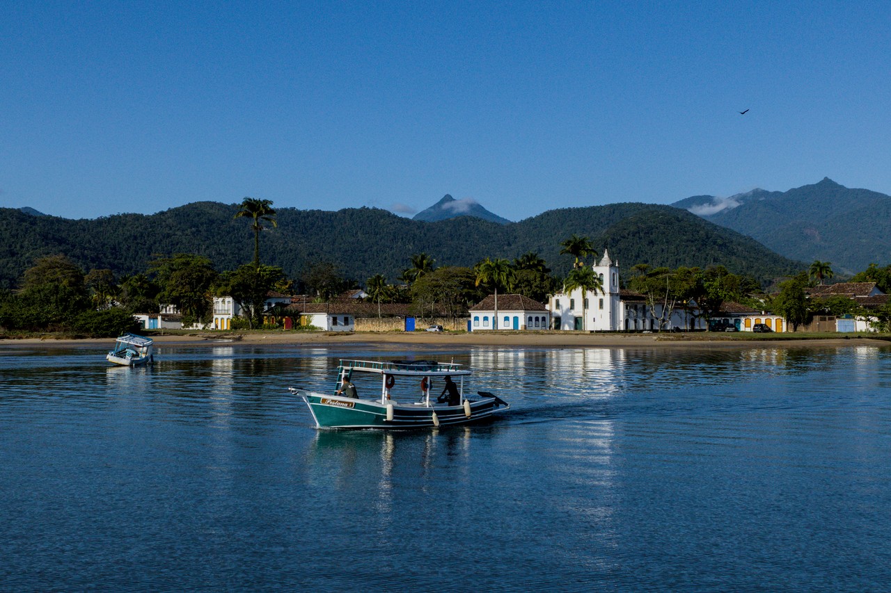 Serras de Paraty. Barco cruza rio em primeiro plano. Ao fundo, igreja branca e serras. Ave corta o céu azul.