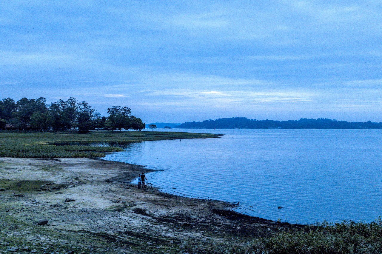 Praia de água doce onde pessoas aparecem de pé à margem ao lado de bicileta; ao fundo, terrenos alagados e colinas vegetadas.