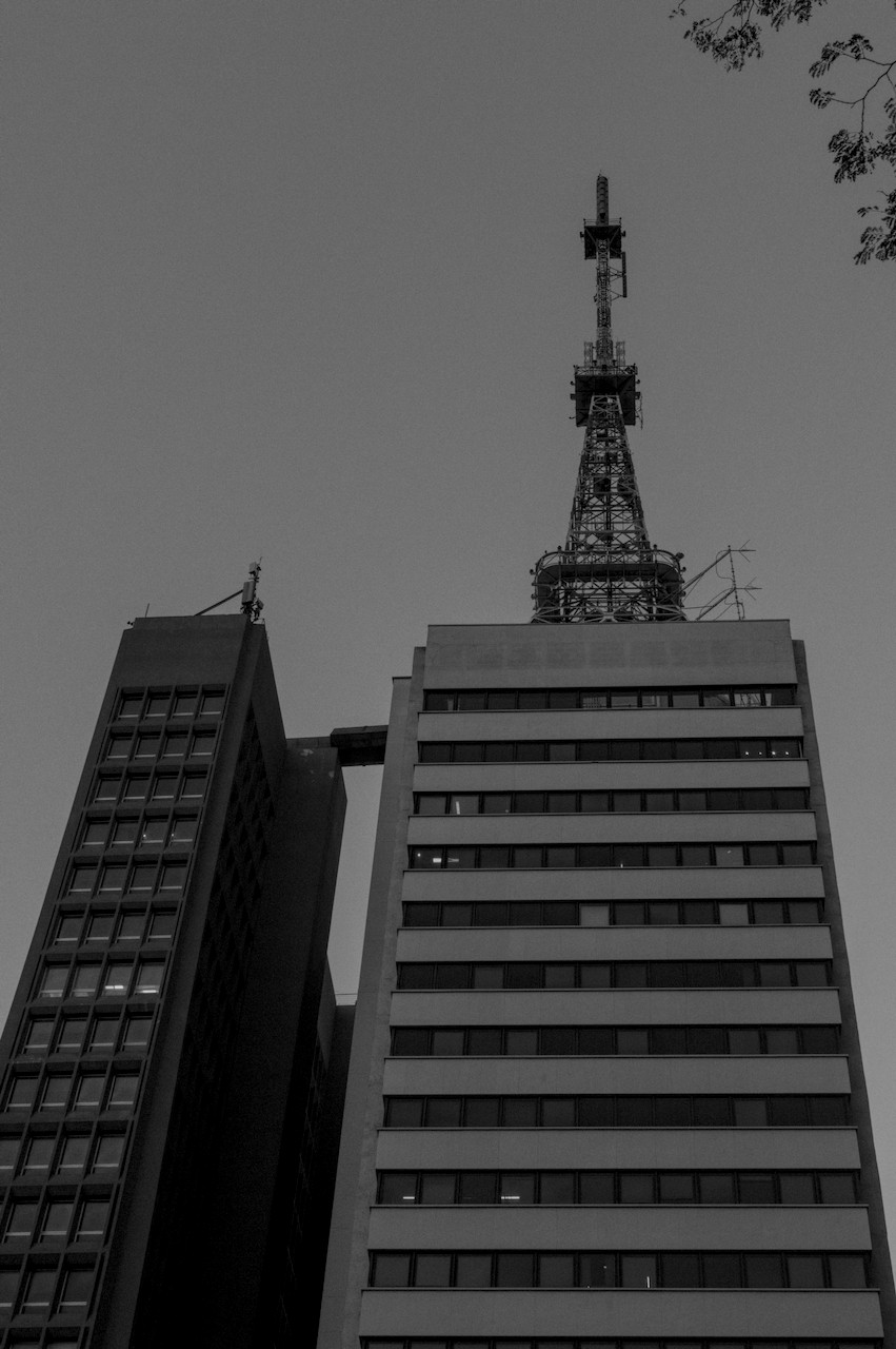 Contra-plongée de dois prédios conectados por passarela, sobre o da direita torre metálica.