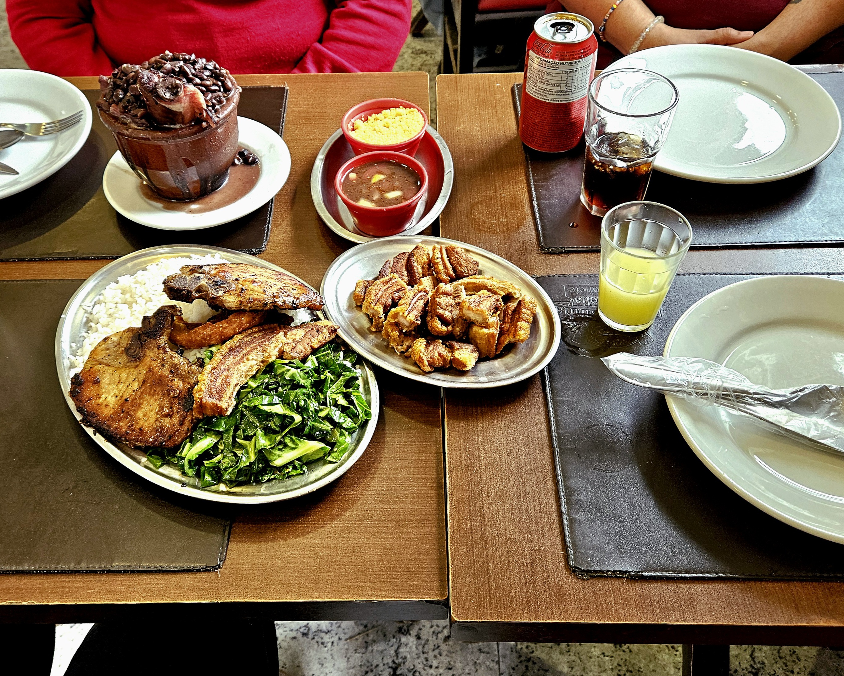 Mesas unidas com pratos, porção de torresmos, cuia de barro com feijoada, prato de arrz com bisteca, couve e torresmo, potinhos de farofa e molho; caipirinha e refrigerante americano; braços de duas pessoas sentadas.
