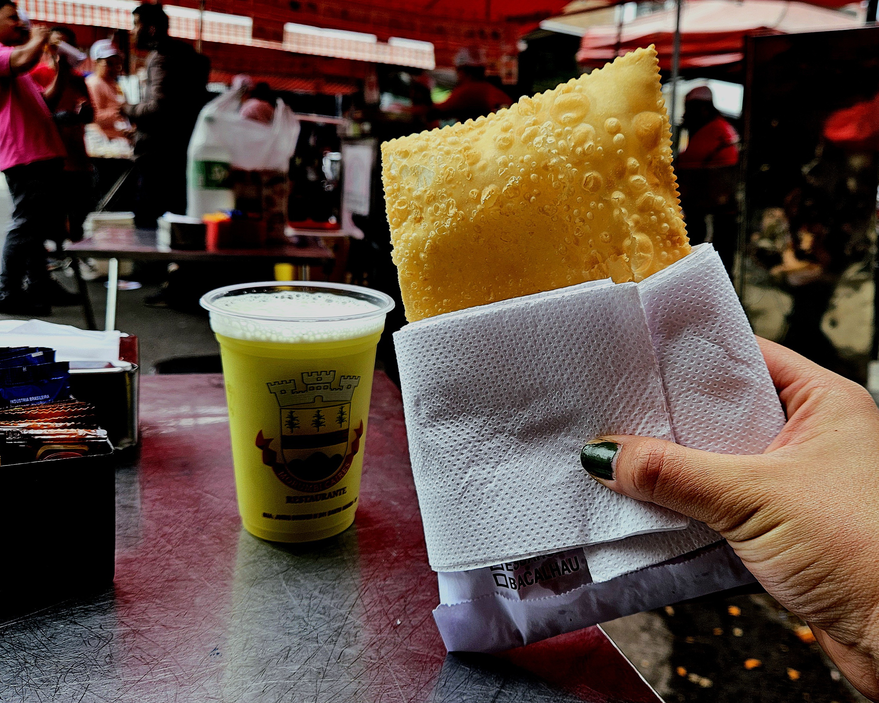 Mesa com pastel em primeiro plano, copo de caldo-de-cana em segundo e feira ao fundo.