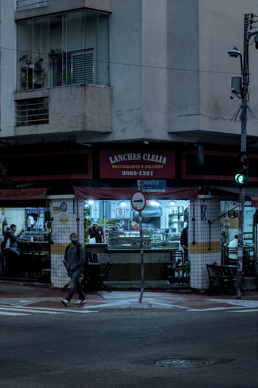 Homem atravessa a rua diante de boteco de esquina iluminado sob o escuro da madrugada.