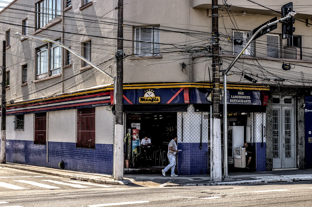 Boteco de esquina sob Sol duro do meio-dia; pessoa caminha ao Sol e outra consome sentada à porta.